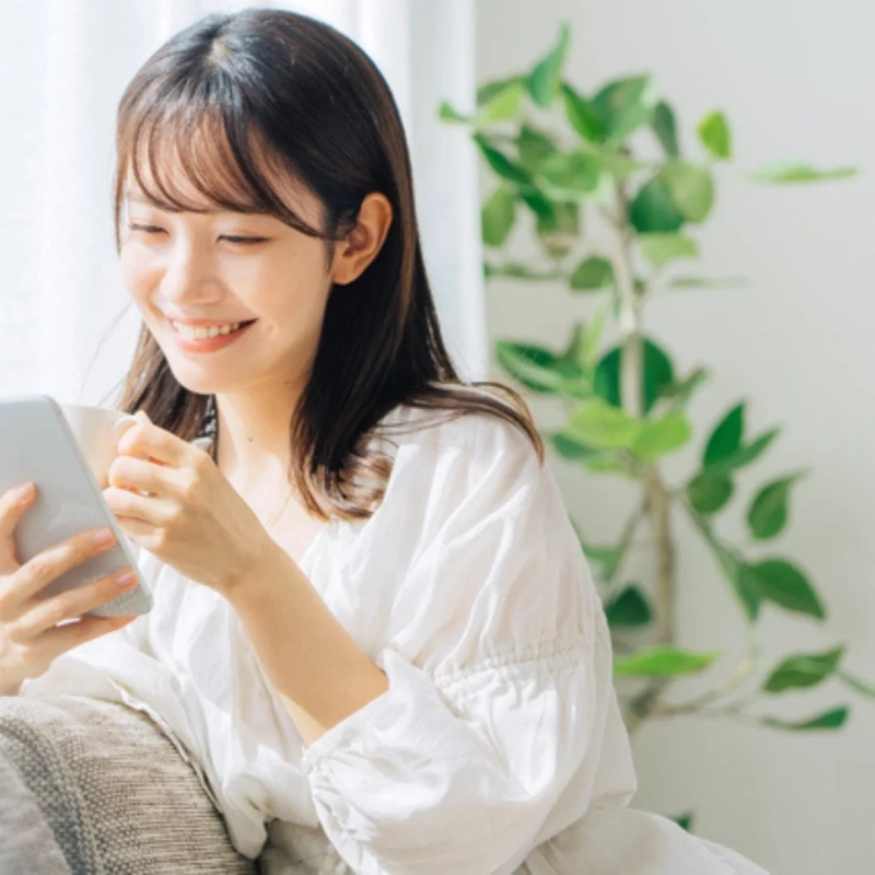 若い女性が自宅でリラックスしながらスマートフォンを操作し、笑顔でコーヒーを飲んでいる様子