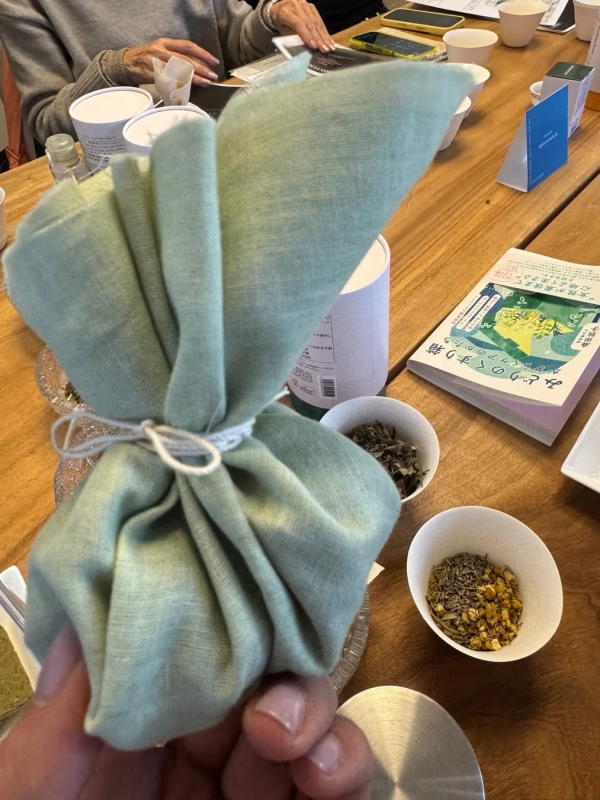ハーブのワークショップらしき場所で、手元に緑色の布で包まれた小包を持ち、テーブルにハーブや本が並んでいる画像