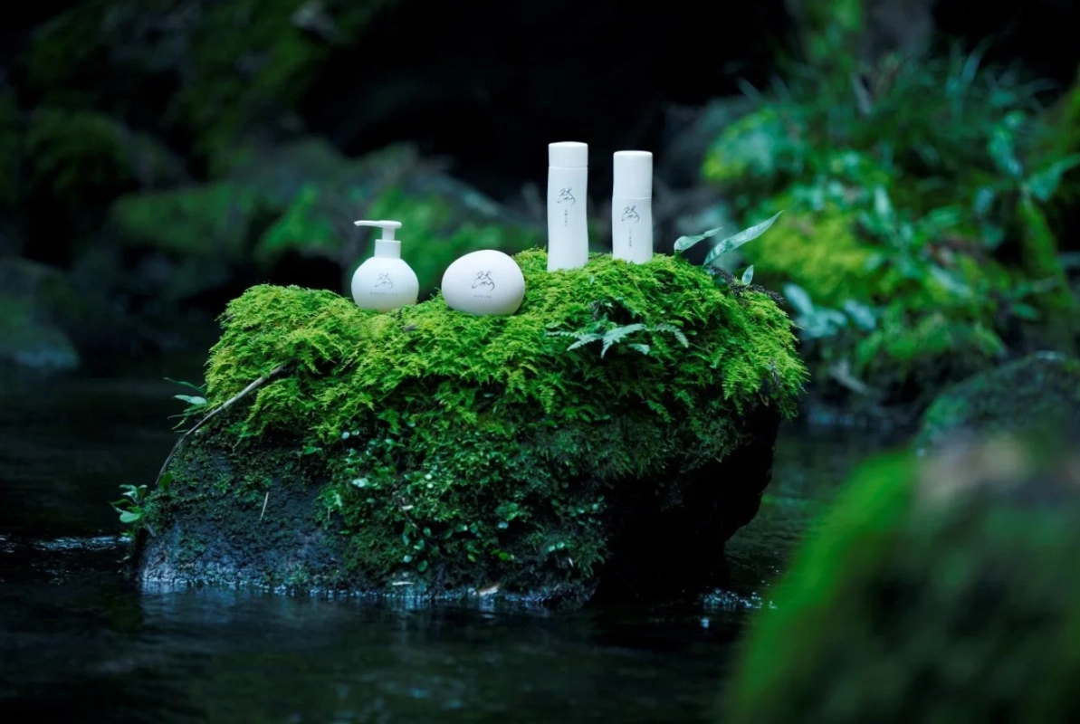 苔むした岩の上に白いスキンケア製品のボトルが置かれ、清らかな水が流れる森の中の風景