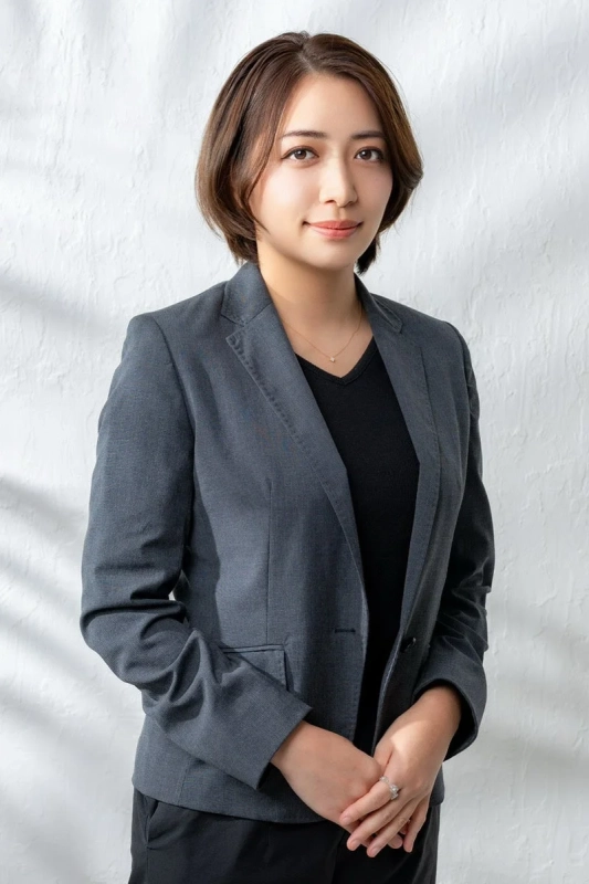 グレーのジャケットと黒いインナーを着用したショートヘアの若い女性のポートレート。優しい笑顔でカメラを見つめており、プロフェッショナルな印象を与えます。