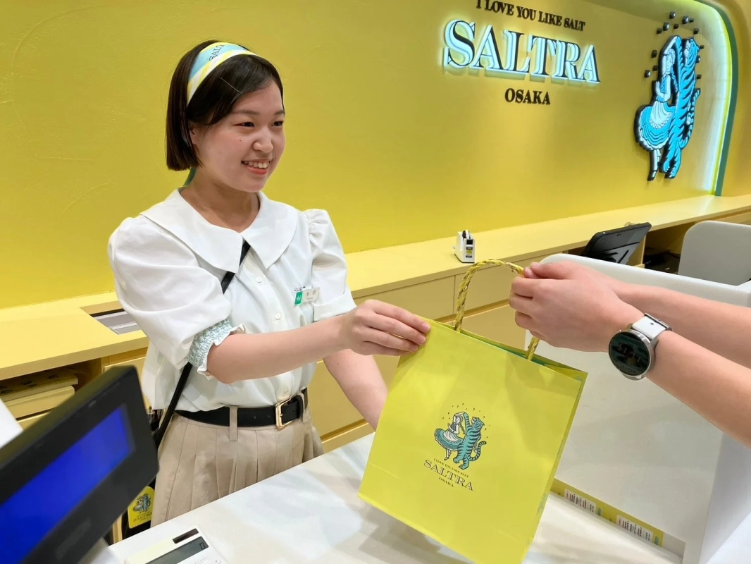 黄色の壁の店舗で、笑顔の女性店員がお客様に「SALT」とロゴの入った黄色の紙袋を手渡している様子。
